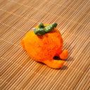 Small sculpture of an orange with 2 green leaves, and a cute cartoon face with purple eyes looking to the side and a simple smile. It lays on its "stomach" with its feet up and head resting in its hands.