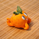 Small sculpture of an orange with 2 green leaves, and a cute cartoon face with purple eyes looking to the side and a simple smile. It lays on its "stomach" with its feet up and head resting in its hands.