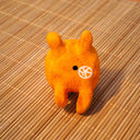 Small sculpture of an orange with 2 green leaves, and a cute cartoon face with purple eyes looking to the side and a simple smile. It lays on its "stomach" with its feet up and head resting in its hands.