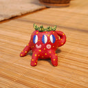 Small ceramic sculpture of a strawberry, with green leaves, yellow spots for its seeds and 3 eyes. It has 2 mouths, one is smiling and the other is a surprised "o". It sits with one arm balancing it and the other behind its head.
