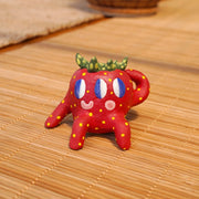 Small ceramic sculpture of a strawberry, with green leaves, yellow spots for its seeds and 3 eyes. It has 2 mouths, one is smiling and the other is a surprised "o". It sits with one arm balancing it and the other behind its head.