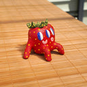 Small ceramic sculpture of a strawberry, with green leaves, yellow spots for its seeds and 3 eyes. It has 2 mouths, one is smiling and the other is a surprised "o". It sits with one arm balancing it and the other behind its head.