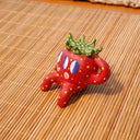 Small ceramic sculpture of a strawberry, with green leaves, yellow spots for its seeds and 3 eyes. It has 2 mouths, one is smiling and the other is a surprised "o". It sits with one arm balancing it and the other behind its head.