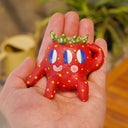 Small ceramic sculpture of a strawberry, with green leaves, yellow spots for its seeds and 3 eyes. It has 2 mouths, one is smiling and the other is a surprised "o". It sits with one arm balancing it and the other behind its head.