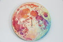 Painting on a circular canvas of a planet with orange and red craters and blue and green shading. It has a simple closed eye expression and a single sparkle shaped tear comes out of the right eye.
