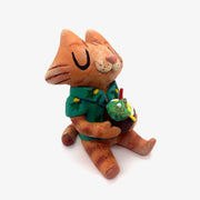 Sculpture of a cartoon style orange tabby cat, sitting with its legs out in front and eyes closed. It holds a pińa colada in its paws and wears a Hawaiian shirt. Viewed from the side.