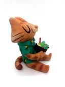 Sculpture of a cartoon style orange tabby cat, sitting with its legs out in front and eyes closed. It holds a pińa colada in its paws and wears a Hawaiian shirt. Viewed from the side.