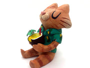 Sculpture of a cartoon style orange tabby cat, sitting with its legs out in front and eyes closed. It holds a pińa colada in its paws and wears a Hawaiian shirt. Viewed from the side.