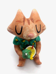 Sculpture of a cartoon style orange tabby cat, sitting with its legs out in front and eyes closed. It holds a pińa colada in its paws and wears a Hawaiian shirt. Viewed from above.