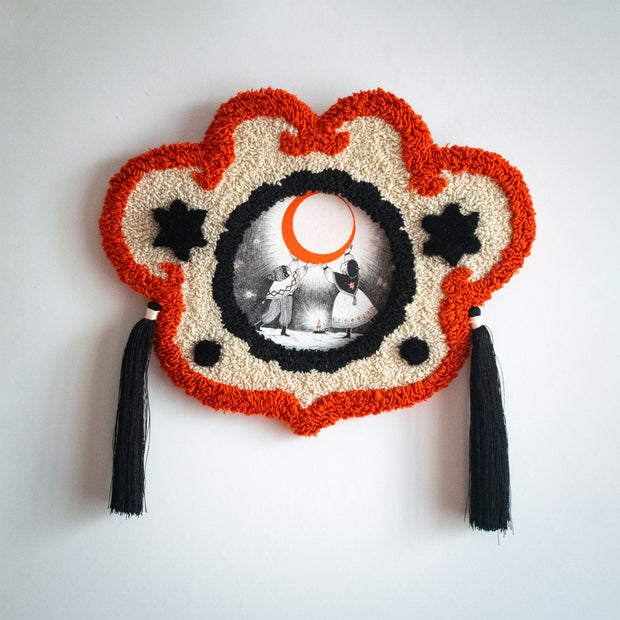 Artwork in ornate embroidered frame with tassels. Artwork is an Ink illustration of a boy and a girl dressed for winter, standing in the snow and holding up a giant orange crescent moon. A candle is on the ground between them.