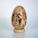 A carved wooden decorative egg sculpture with detailed hand-painted scenes of animals and trees, displayed on a small round wooden stand against a light gray background.