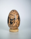 A carved wooden decorative egg sculpture with detailed hand-painted scenes of animals and trees, displayed on a small round wooden stand against a light gray background.
