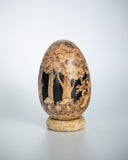 A carved wooden decorative egg sculpture with detailed hand-painted scenes of animals and trees, displayed on a small round wooden stand against a light gray background.