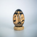A decorative wooden egg with a glossy black finish and hand-painted animal and floral motifs, displayed on a small round cork stand against a light gray gradient background.