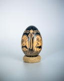 A decorative wooden egg with a glossy black finish and hand-painted animal and floral motifs, displayed on a small round cork stand against a light gray gradient background.
