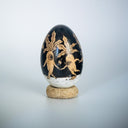 A decorative wooden egg with a glossy black ink illustration of botanical and animal motifs, resting on a small round cork base against a light gray background.
