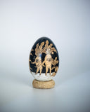 A decorative wooden egg with a glossy black ink illustration of botanical and animal motifs, resting on a small round cork base against a light gray background.