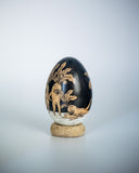 A decorative wooden egg with a glossy black ink illustration of botanical and animal motifs, resting on a small round cork base against a light gray background.