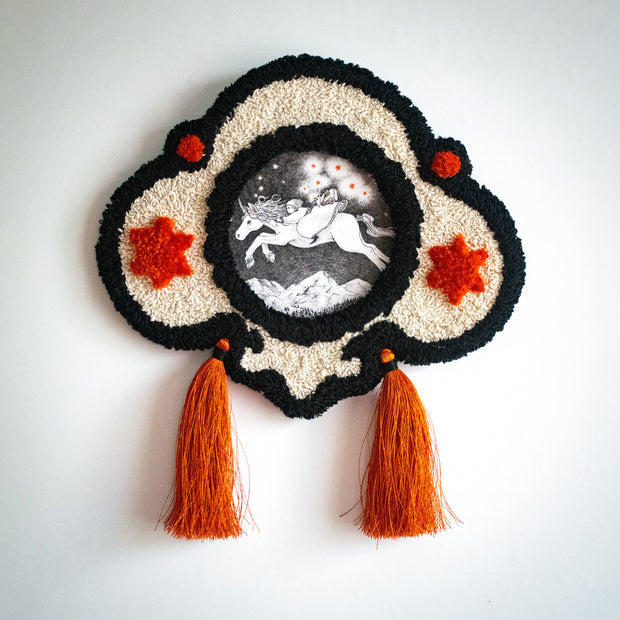 Artwork with an ornate embroidered frame with long tassels. Artwork is an Ink illustration of a girl riding atop of a unicorn, which jumps through the night sky. Orange stars trail behind them, coming out of the girl's side pouch.