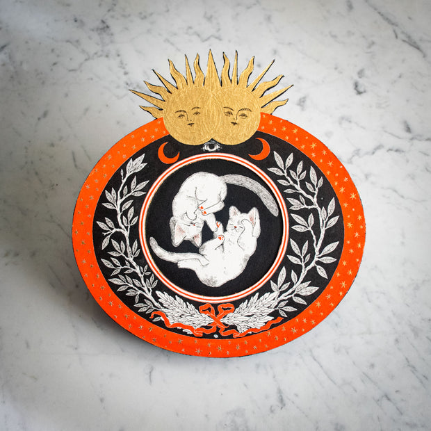 Black and orange ink illustration of 2 cats, floating opposite one another like yin & yang. They are in a circle surrounded by floral patternings. 2 gold suns are at the top of the piece.