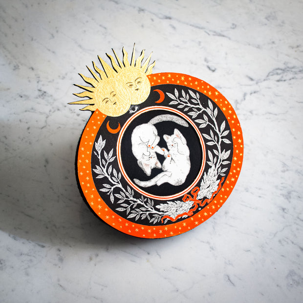 Black and orange ink illustration of 2 cats, floating opposite one another like yin & yang. They are in a circle surrounded by floral patternings. 2 gold suns are at the top of the piece.