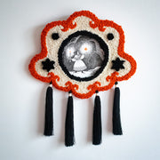 Artwork with an ornate, hand embroidered frame with long black tassels. The artwork is an Ink illustration of a girl holding a candlestick and holding out her other hand, where an orange star hovers above it. She stands in a forest in the snow.