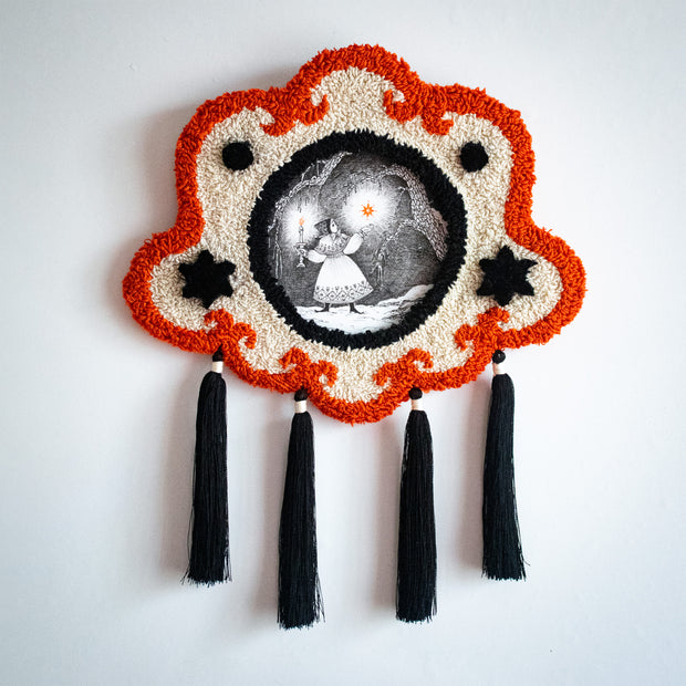 Artwork with an ornate, hand embroidered frame with long black tassels. The artwork is an Ink illustration of a girl holding a candlestick and holding out her other hand, where an orange star hovers above it. She stands in a forest in the snow.
