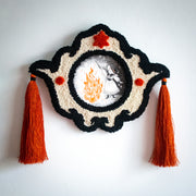 Decorative wall piece with tassels embroidered detailing and with an artwork within it - Illustration of a witch wearing a dress and a hat and holding a wand, conjuring a very large fire, which casts the surroundings into darkness.