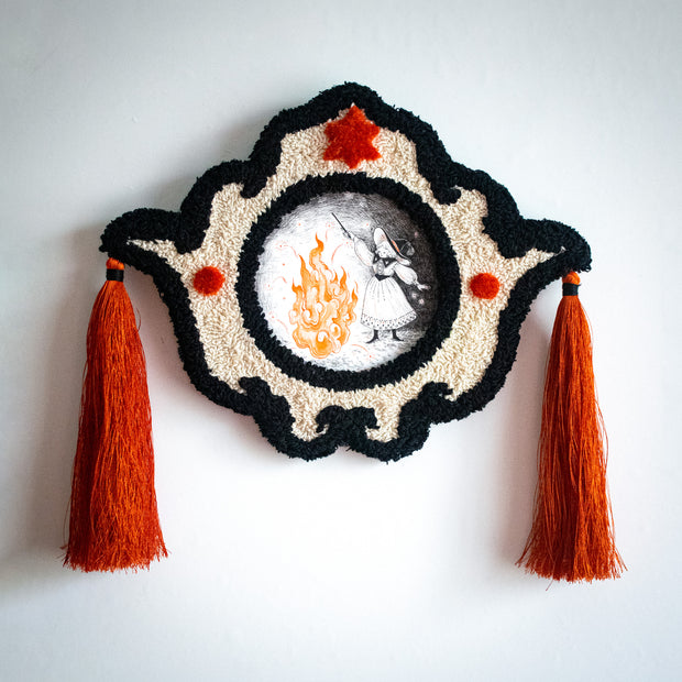 Decorative wall piece with tassels embroidered detailing and with an artwork within it - Illustration of a witch wearing a dress and a hat and holding a wand, conjuring a very large fire, which casts the surroundings into darkness.