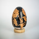 A decorative wooden egg with black illustrations of floral and animal illustrations, displayed upright on a small cork-like base against a light gray studio background.
