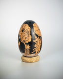 A decorative wooden egg with black illustrations of floral and animal illustrations, displayed upright on a small cork-like base against a light gray studio background.