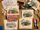 A styled flat-lay of cartoon art prints and sketching tools on a brown cardboard surface. Multiple printed sheets show a group of cute animal characters gathered around a green table, with some prints in black ink, one in muted pink, and one in cream.