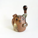 Ceramic sculpture of a cute, brown dog with its paws on its cheek and a rounded bottom without legs. It has green textured spots. It holds an upside down wooden awl between its arm.