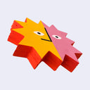 Die cut wooden painting in the shape of a multi pointed sun,, half yellow and half pink with only eyes and a straight nose.