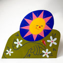 A colorful folk-art style wooden diecut sculpture of a smiling yellow sun with pink rays over an olive green hill-like body with white flowers and a small red bird, photographed against a plain light background.