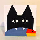 Painting on wood panel of a black cat, with a very simplistic silhouette, seen only from the chin up. It holds a tricolor fish in its mouth with its 2 fangs showing.
