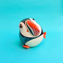 Ceramic sculpture of a puffin with a very rounded, chubby body. It sits with one of its legs lifted.