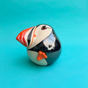 Ceramic sculpture of a puffin with a very rounded, chubby body. It sits with one of its legs lifted.
