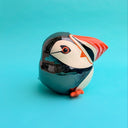 Ceramic sculpture of a puffin with a very rounded, chubby body. It sits with one of its legs lifted.