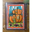 Artwork featuring a whimsical illustration of orange pumpkins shaped like houses with a green door, mushrooms, and a spider, surrounded by an orange wavy border and housed in a clear protective sleeve, placed on a wooden surface.
