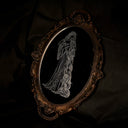 An ornate oval mirror with a dark bronze-brown carved frame, featuring a black reflective surface with a white line-art engraving of a woman with very long, braided hair and a long billowy dress, set against a dark draped fabric background.