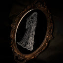 An ornate oval mirror with a dark bronze-brown carved frame, featuring a black reflective surface with a white line-art engraving of a woman with very long, braided hair and a long billowy dress, set against a dark draped fabric background.