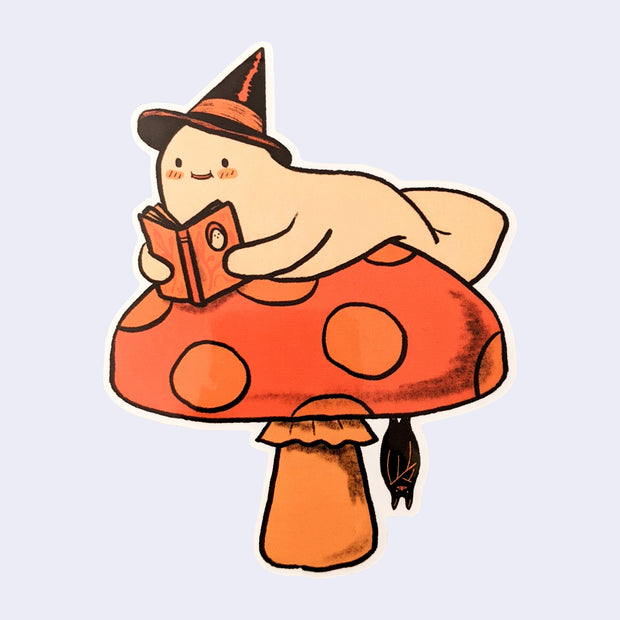 Die cut sticker of a ghost reading a book and resting on a mushroom, which a bat hangs from the underside.
