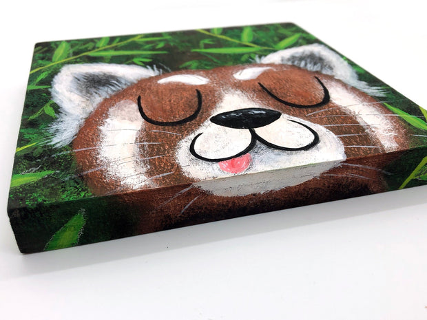 Illustrative style painting of a cute, red panda with large closed eyes and a smiling tongue out expression. Only its head is visible but it takes up most of the surface, with bamboo behind it.