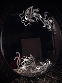 A round black mirror featuring white etched illustrations of 2 koi fish with lily pads, one atop and one at the bottom, set against a dark quilted wall and dark marble surface.