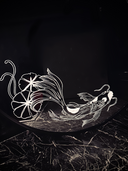A round black mirror featuring white etched illustrations of 2 koi fish with lily pads, one atop and one at the bottom, set against a dark quilted wall and dark marble surface.