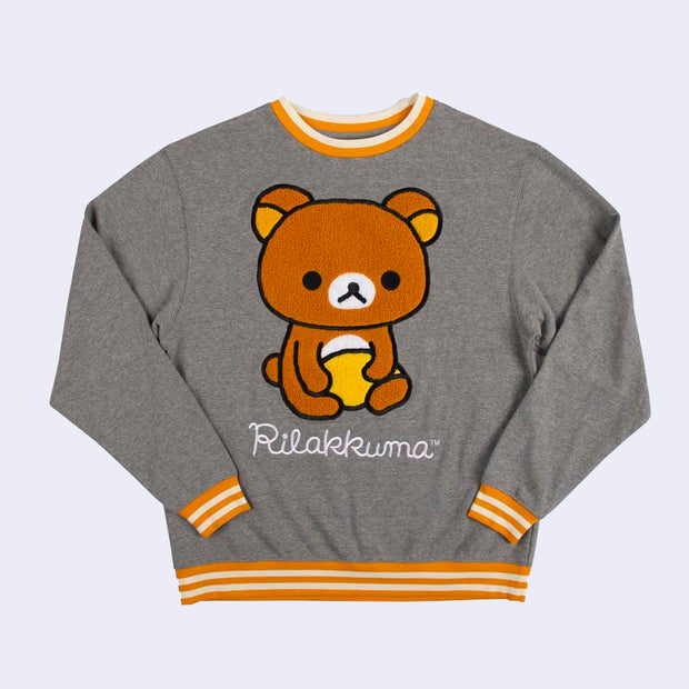 Gray pull over sweater with a large plush graphic of Rilakumma, with "Rilakkuma" written below in cursive. The collar, sleeve holes and bottom of the sweater have an orange and white striped binding.