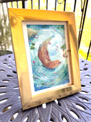 Framed artwork of a brown otter in water with gold frame on a metal table.