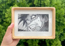 Graphite illustration of Gaara from Naruto, a close up of his face as part of it is cracked off to reveal sand underneath. He has a scary expression with gritted teeth and wide eyes. Piece is in a wooden frame with rounded edges.