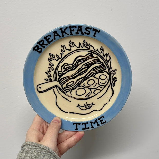 Hand holding a ceramic plate with 'Breakfast Time' text and food illustration on a plain background